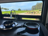 RV rental kitchen view: A neat setup with a pan, blue dishes, and sink, overlooking a beautiful green landscape and ocean.