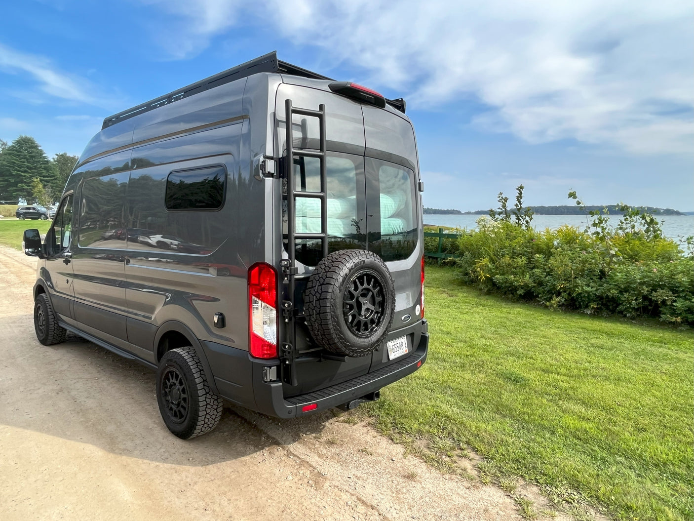 Modern gray Ford Transit camper van with roof rack and rear spare, ready for an RV rental adventure near the water.