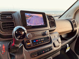 This RV rental interior shows the dashboard, gear shifter, and controls, with a backup camera displaying a beachside view.