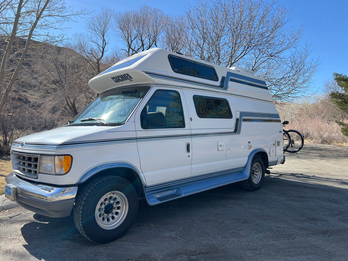 Explore in this white Ford Econoline Coachmen camper van, an ideal RV rental, parked with a bike rack & mountain backdrop.