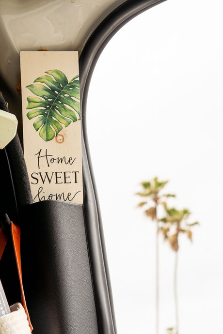 Home sweet home in your RV rental! A decorative sign with a Monstera leaf is seen inside, with palm trees in the blurry background.
