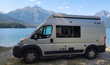 Scenic RV rental camper van parked by a serene lake with majestic mountains and forests in the background. Perfect for nature travel.