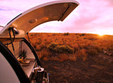 RV rental camping: Teardrop camper's outdoor kitchen with brewing coffee, set against a vibrant sunset over arid land.