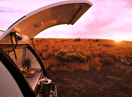 RV rental camping: Teardrop camper's outdoor kitchen with brewing coffee, set against a vibrant sunset over arid land.