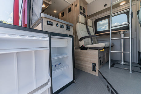 RV rental interior: mini-fridge with water, modern control panel, passenger seating, and table with ladder access.