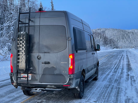 An RV rental, a gray Mercedes Sprinter van, parked on an icy Alaska road amid snowy trees, ready for winter adventure.