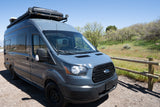 Custom grey Ford Transit camper van for rv rental, parked outdoors on a sunny day next to a wooden fence with green trees.