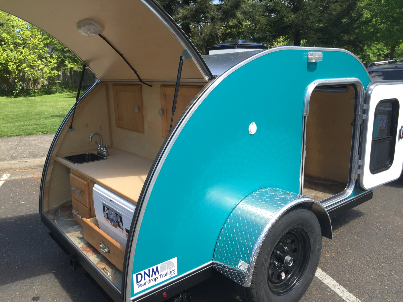 Explore this turquoise teardrop trailer, perfect for RV rental, showcasing its open rear kitchenette & side sleeping area access.