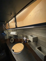 Compact RV rental kitchen area featuring a dark countertop, sink with cutting board, wall storage, and lighted overhead cabinets.
