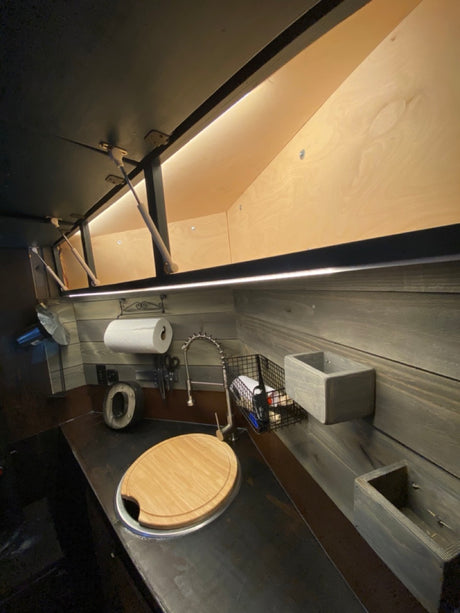 Compact RV rental kitchen area featuring a dark countertop, sink with cutting board, wall storage, and lighted overhead cabinets.