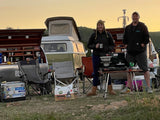 A couple smiles, cooking dinner with an RV rental campervan & truck setup, chairs, and cooler at dusk.