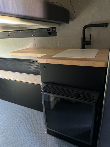 RV rental interior: Modern compact kitchen with black faucet, wood counter, fridge, overhead bed, and patterned floor.