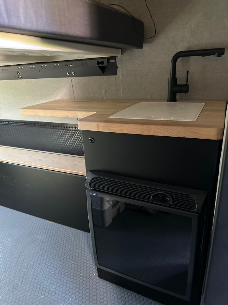 RV rental interior: Modern compact kitchen with black faucet, wood counter, fridge, overhead bed, and patterned floor. — photo 11