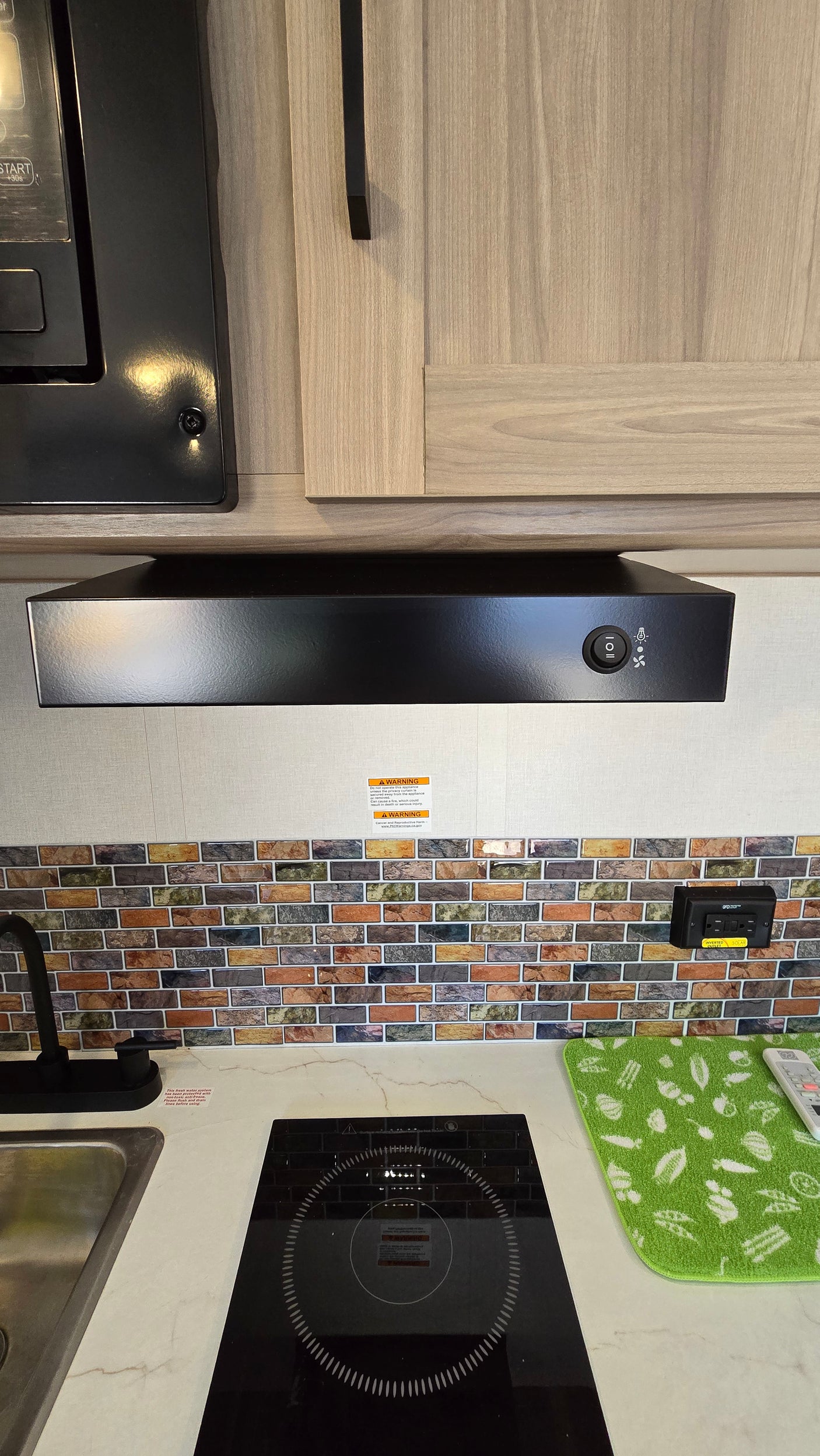 RV rental kitchen: modern induction cooktop, sink, range hood, and colorful tile backsplash under light wood cabinets.