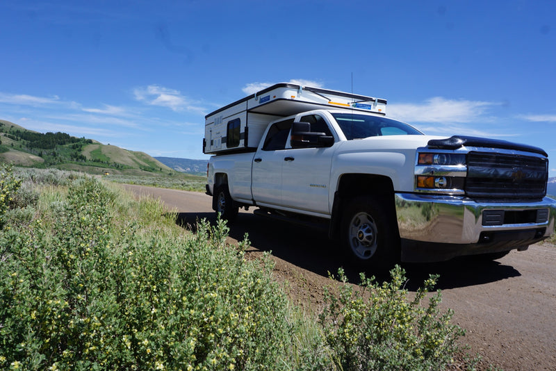  parked on a dirt road by snow-capped mountains.|Wyoming's Grand Tetons frame this white truck camper RV rental on a dirt road. Perfect for mountain adventures.|White Chevy Silverado 2500 HD truck with a Four Wheel truck camper on a dirt mountain road. Perfect for an rv rental trip.|Front view of a white pickup truck with a pop-up RV camper — photo 2