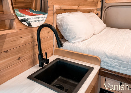 RV rental interior featuring wood paneling, a circular mirror, a compact black sink/faucet, & cozy bed.