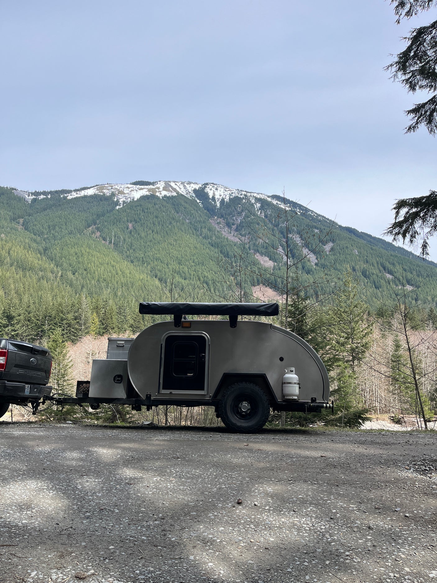 Scenic RV rental adventure with a truck and teardrop trailer parked amidst snowy mountains and a dense forest.