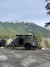 Scenic RV rental adventure with a truck and teardrop trailer parked amidst snowy mountains and a dense forest.