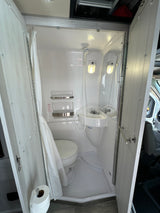 A compact, all-in-one bathroom in an RV rental, featuring a toilet, sink, and shower with curtain and non-slip floor.