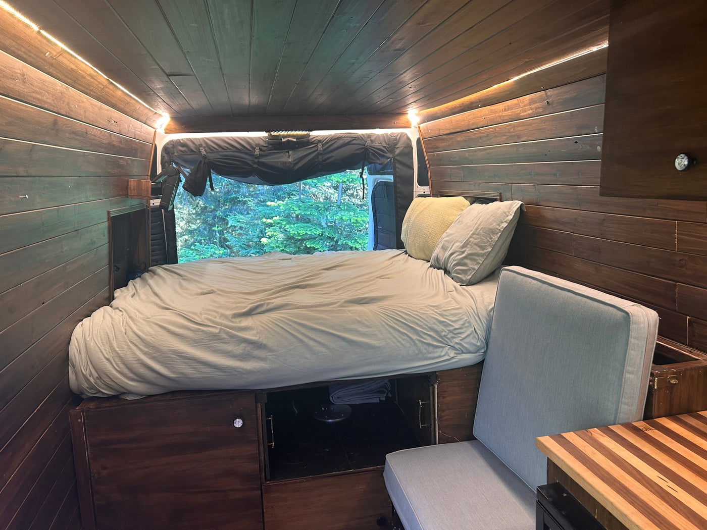 Cozy RV rental interior with wood-paneled walls, bed, ambient strip lighting, window view of trees, cushioned seat, & storage. — photo 9