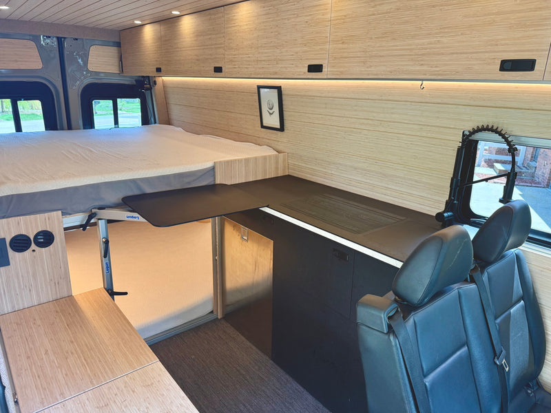 This RV rental interior showcases a raised bed, modern kitchenette with counter, flexible dining table, and passenger seating. — photo 7