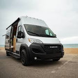 A sleek silver Noova campervan is parked on pavement beside a sandy beach. Ideal for your next RV rental adventure by the sea.