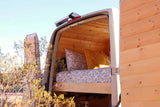An RV rental's cozy custom wood-paneled interior, featuring a bed with patterned pillows, viewed through its open back doors. Ideal for van life.