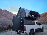 White truck with Alu-Cab camper and open rooftop tent as an RV rental, parked by a scenic river and rocky mountains.