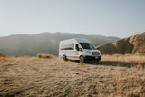 A white RV rental van is parked on a dry, grassy hillside with scenic mountains and a clear sky in the background.