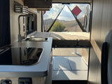 RV rental's cozy interior: kitchen, bed, and scenic hills with a red sign seen through the bug-netted open rear.