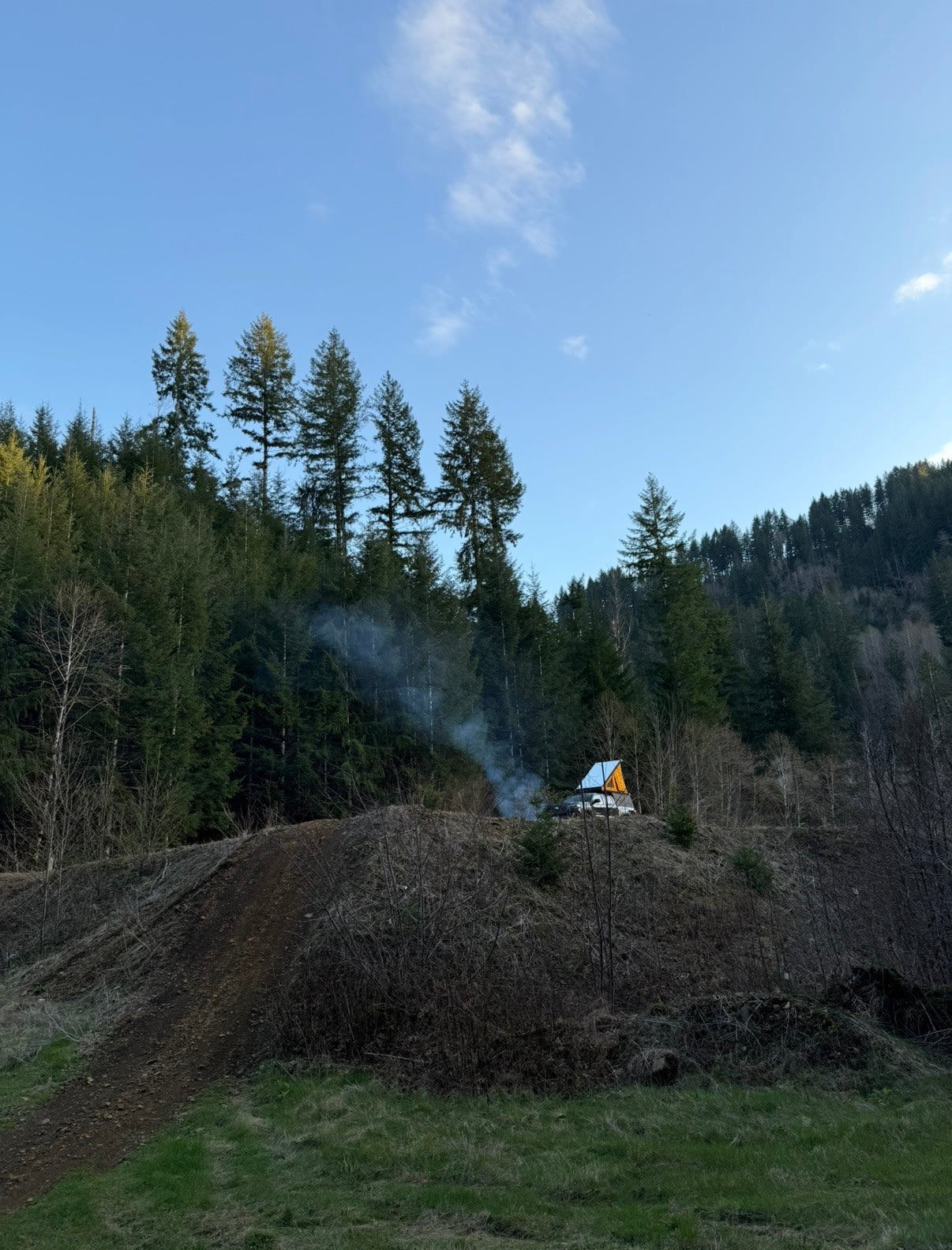An RV rental with a pop-up tent atop a car, nestled in a forest clearing with a smoking campfire under blue skies.