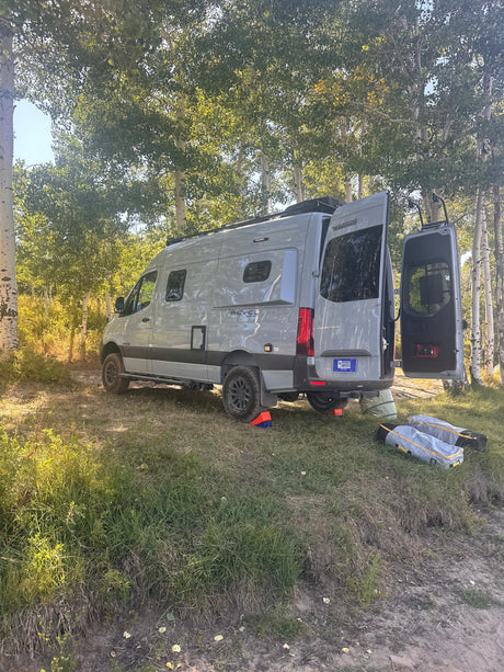 Gray Winnebago Revel rv rental camper van with open rear doors, leveled and ready for camping in an Aspen forest.