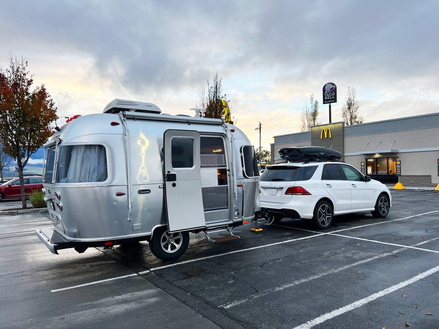 A white SUV tows an Airstream RV rental, parked outside McDonald's & Taco Bell under a cloudy sky. Ready for a road trip!