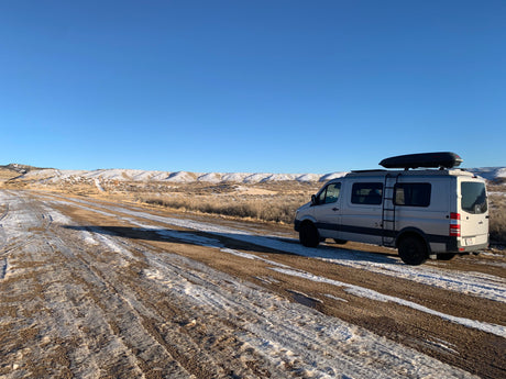 Grey RV rental van parked on a snowy dirt road with distant hills under a clear blue sky, ideal for winter adventures.