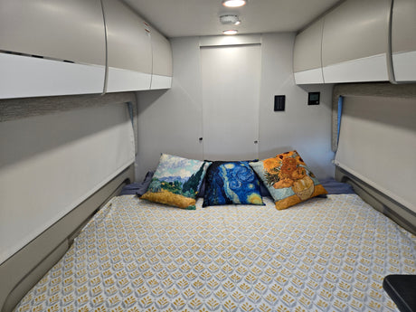 Modern RV rental interior: comfortable bed with artistic pillows, integrated storage, and system controls.