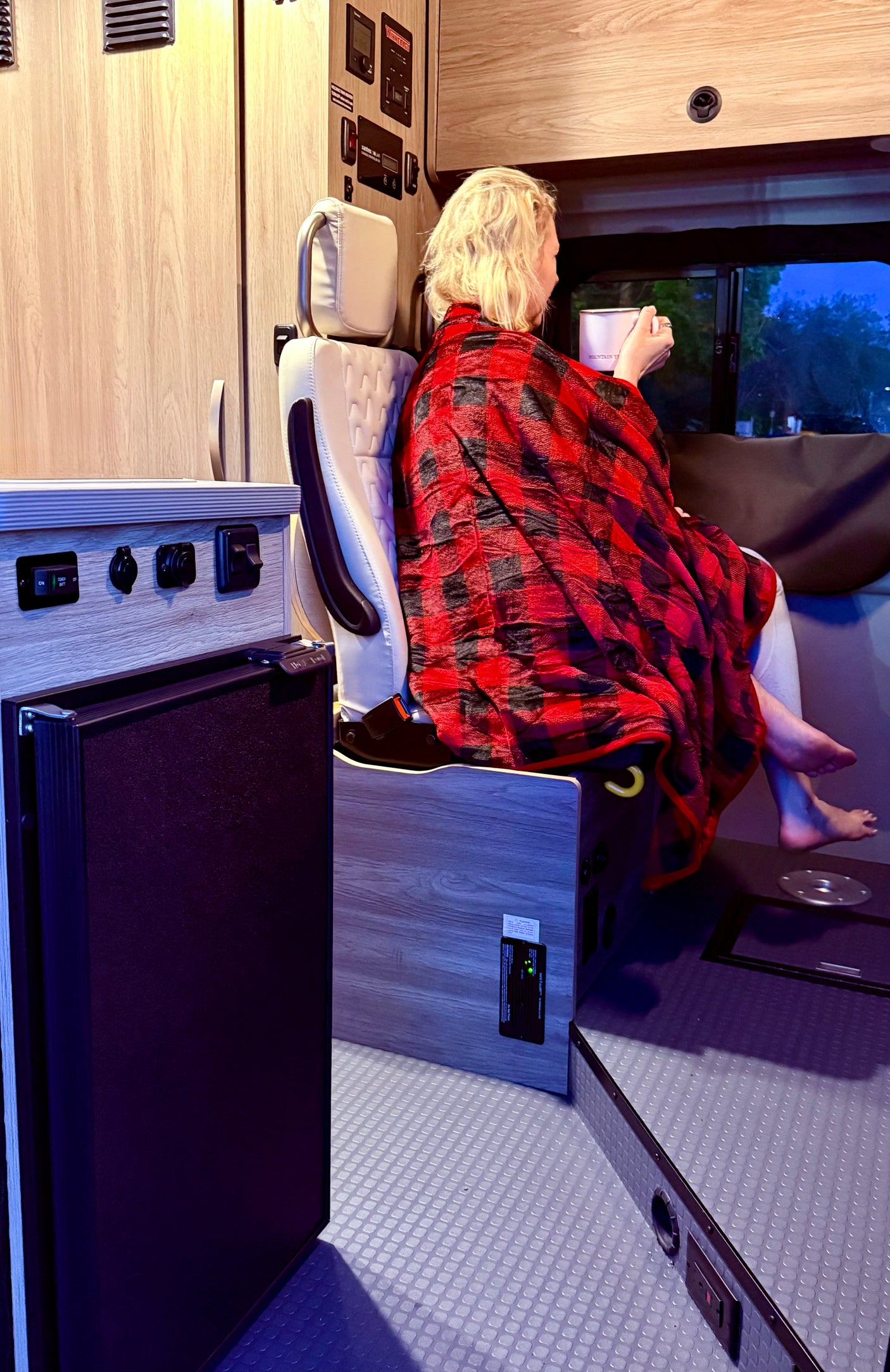 Cozy RV rental evening: person wrapped in plaid blanket sips from a mug, enjoying the view from their camper.
