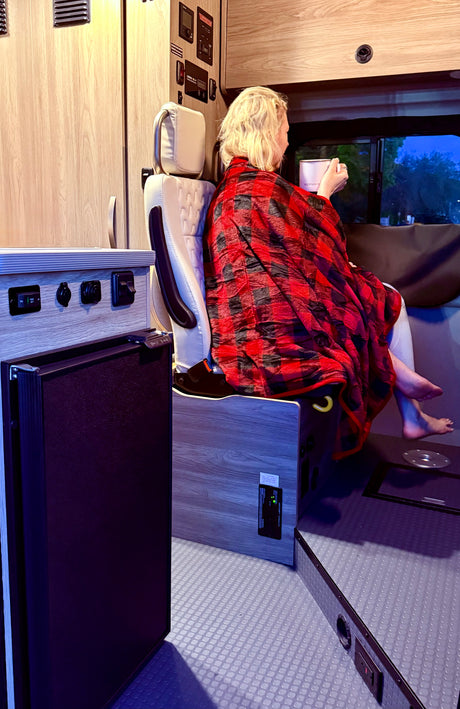 Cozy RV rental evening: person wrapped in plaid blanket sips from a mug, enjoying the view from their camper.