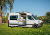 White RV rental camper van parked in a sunny field, side door open revealing a modern van life kitchen for adventure.