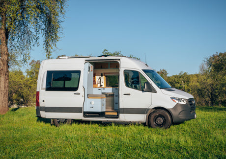 White RV rental camper van parked in a sunny field, side door open revealing a modern van life kitchen for adventure.