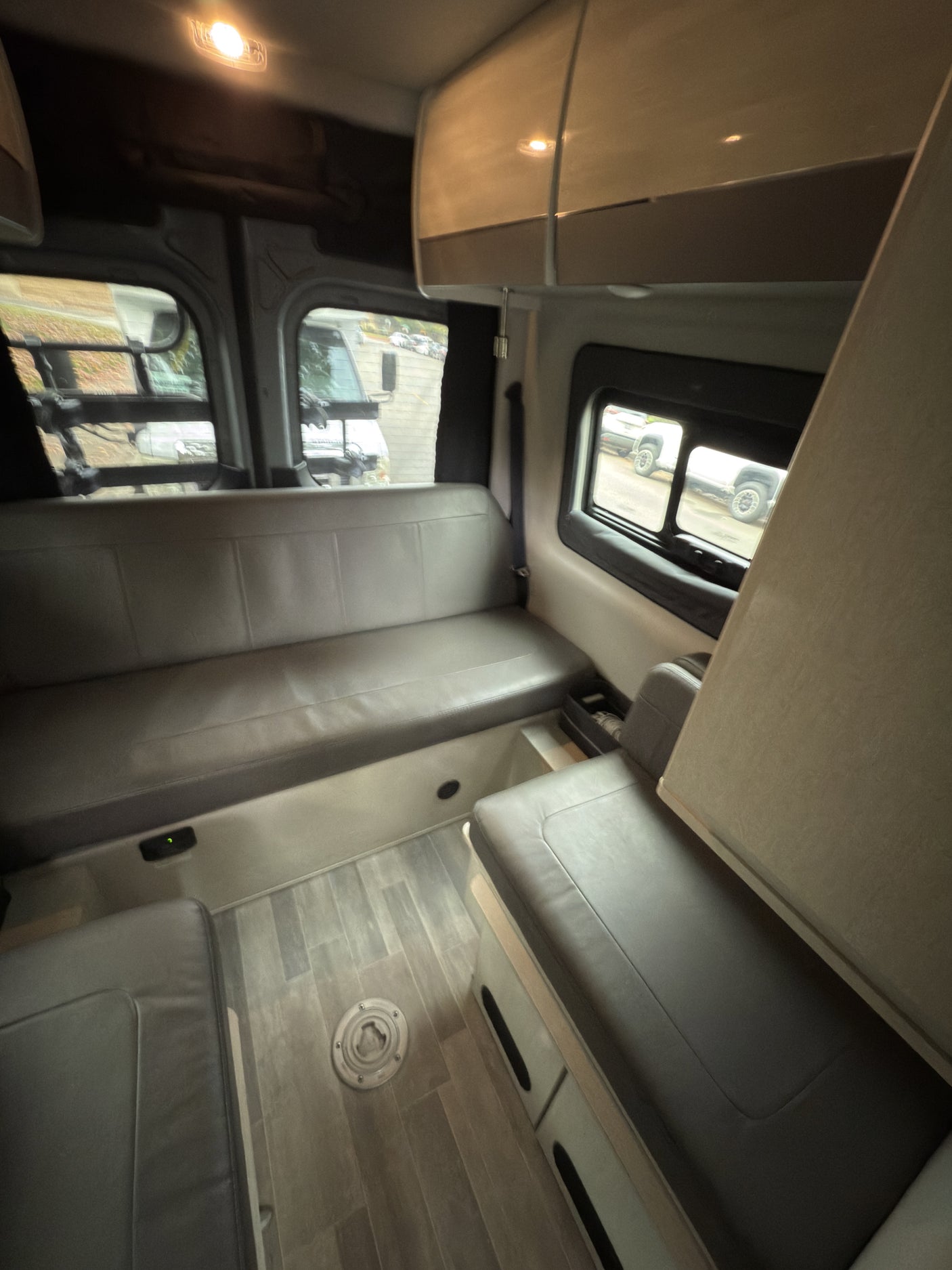 View inside an RV rental: two long grey cushioned benches facing each other, faux wood floor, and side windows.