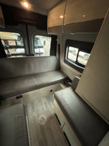 View inside an RV rental: two long grey cushioned benches facing each other, faux wood floor, and side windows.