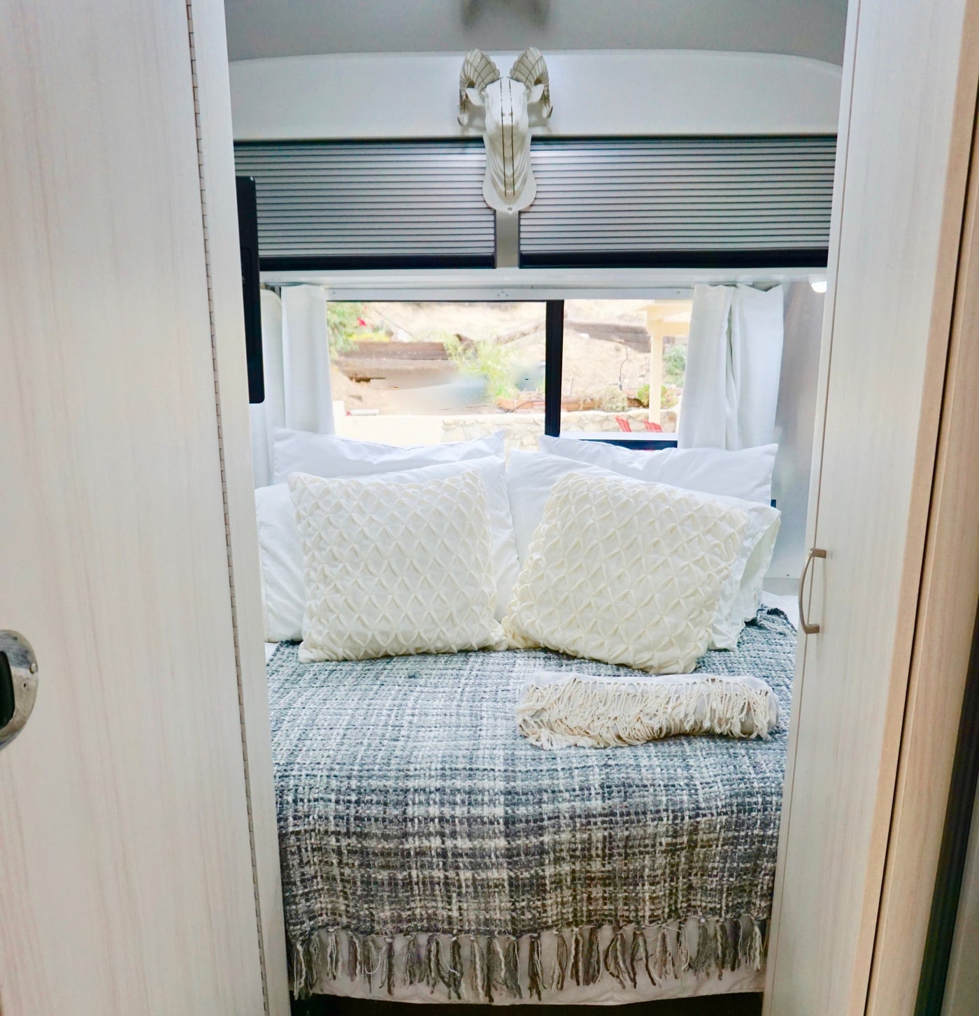 Cozy RV rental bedroom: a bed with white pillows, grey blanket, and ram head decor. Windows reveal a dry outdoor landscape. — photo 11