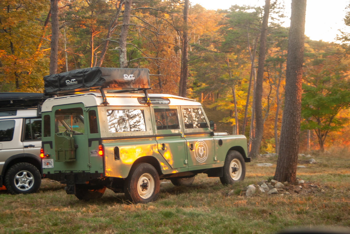 RV rental ready for adventure: Green Land Rover SUV with rooftop tent parked in sunlit autumn woods, another SUV beside it.
