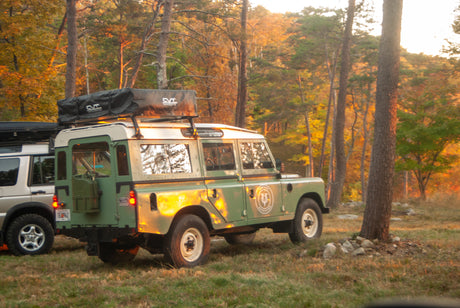 RV rental ready for adventure: Green Land Rover SUV with rooftop tent parked in sunlit autumn woods, another SUV beside it.