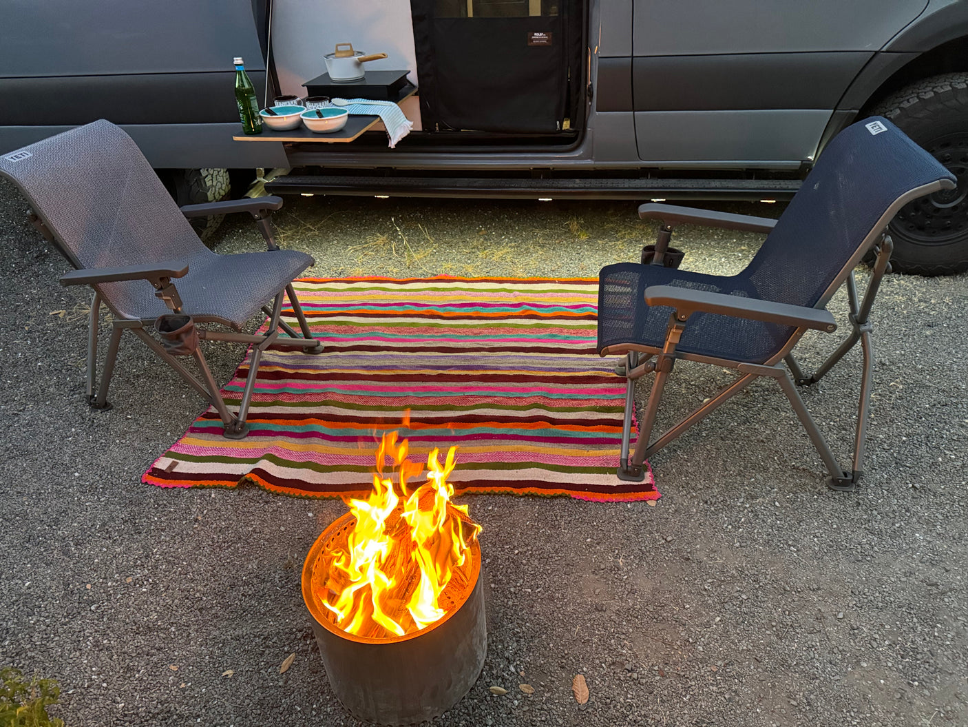 A cozy outdoor scene with a gray RV rental, two camping chairs, a striped rug, and a burning fire pit on gravel.