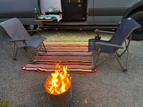 A cozy outdoor scene with a gray RV rental, two camping chairs, a striped rug, and a burning fire pit on gravel.