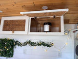 Cozy RV rental interior shows open rattan overhead cabinets with a Ninja blender. Wood ceiling, shiplap walls, and string lights add charm.