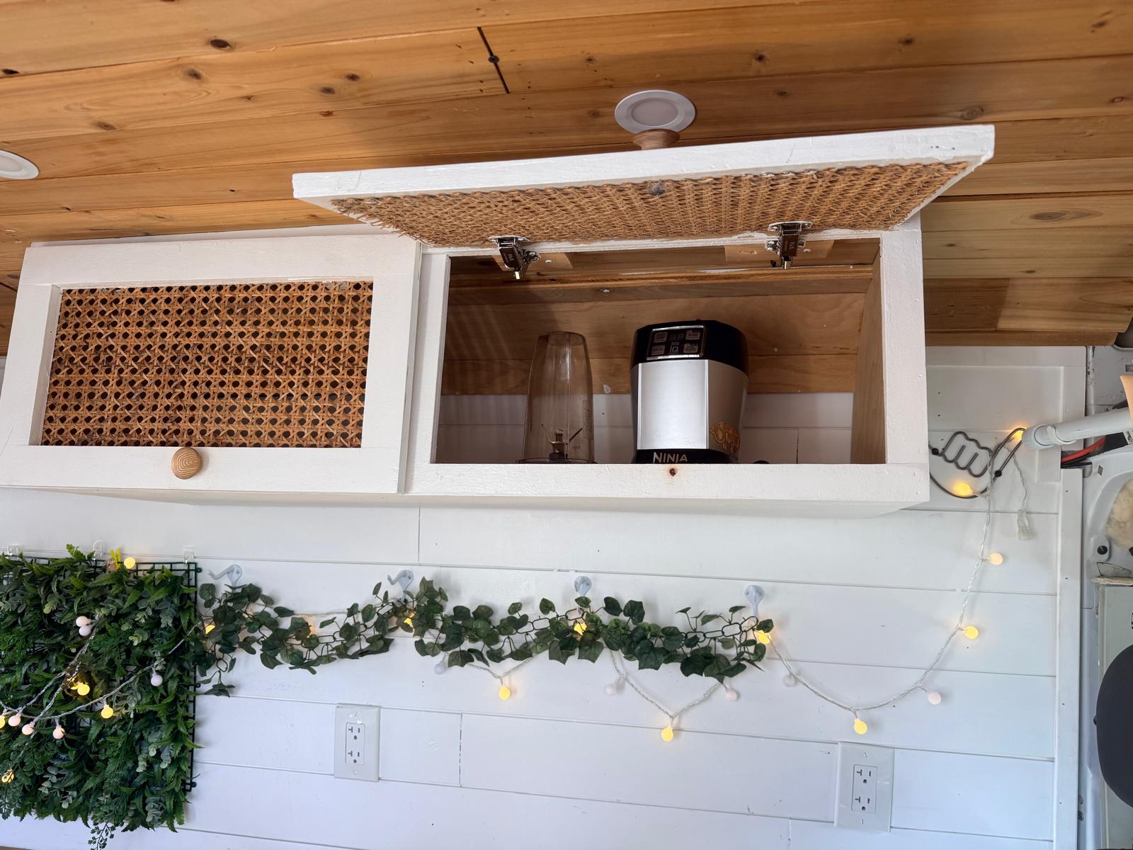 Cozy RV rental interior shows open rattan overhead cabinets with a Ninja blender. Wood ceiling, shiplap walls, and string lights add charm.