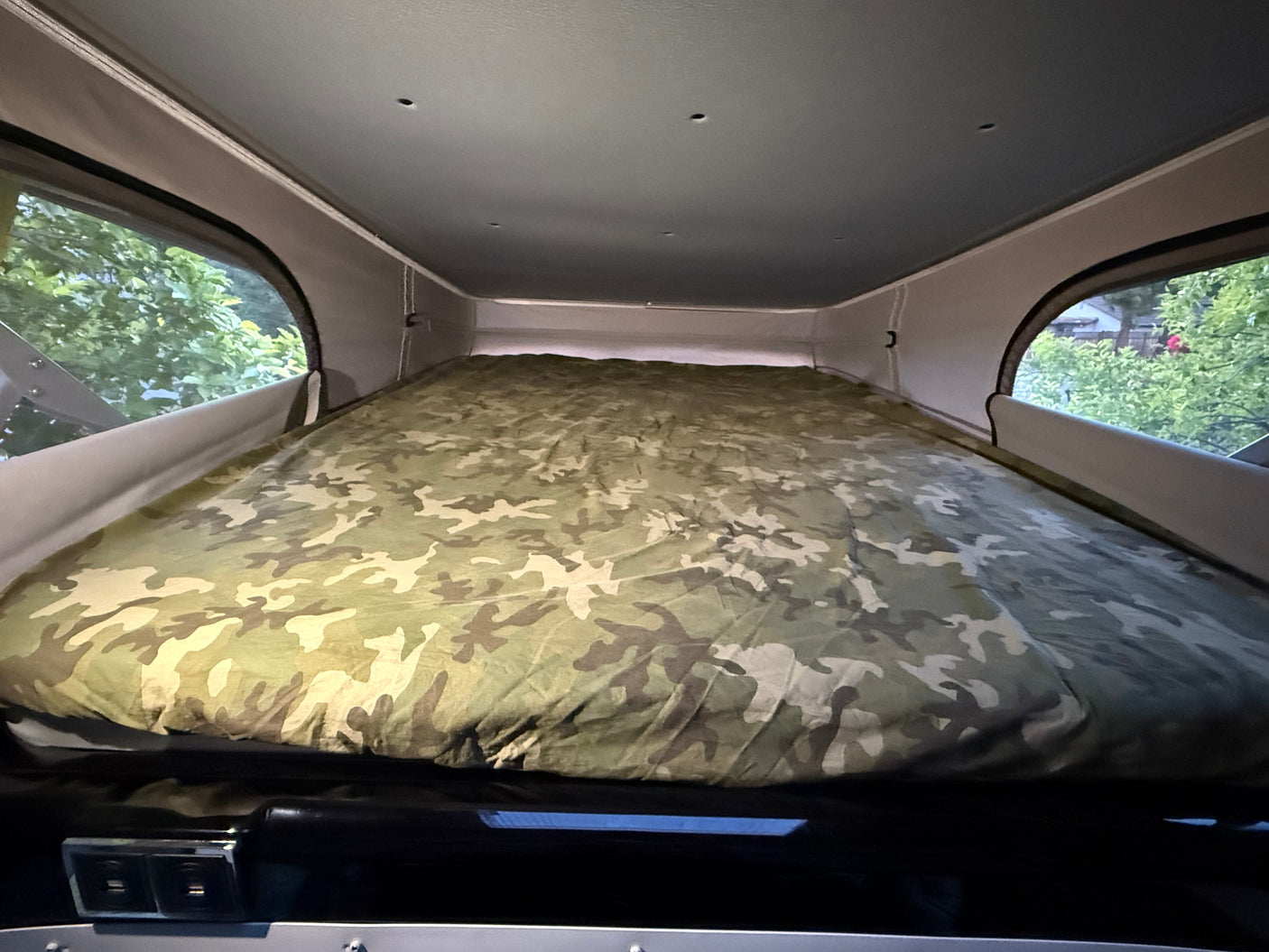 Cozy camo bed inside an RV rental pop-top camper, offering a peaceful view of nature from the windows. Perfect for camping.