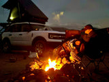 A cozy campfire illuminates a person and a white Ford Bronco Sport with a rooftop tent, ideal for RV rental adventures.